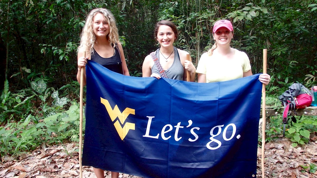 Three female students hold up a West Virginia University “Let’s go.” flag while standing in a jungle-like setting.