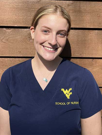 Carley is shown smiling while wearing blue WVU School of Nursing scrubs and standing in front of a wall made of vertical wooden planks.